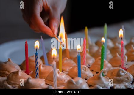 Main tenant des allumettes et allumant des bougies colorées sur le grand gâteau d'anniversaire au chocolat fait maison, décoré de meringues et de crème fouettée. Gros plan Banque D'Images