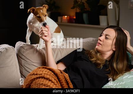 Femme allongée sur le canapé et jouant avec son chien. Femme heureuse se liant avec Jack Russell Terrier chien sur le canapé à la maison. Concept d'affection, de relaxation et d'amour pour animaux de compagnie Banque D'Images