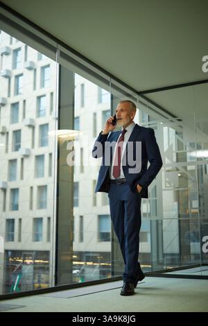 Plan vertical large d'un homme d'affaires caucasien d'âge moyen moderne portant un costume, marchant le long de l'espace de bureau avec la main dans la poche, parlant au téléphone Banque D'Images