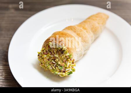 Une corne de pâtisserie brun doré remplie de crème et surmontée de pistaches écrasées, servie sur une assiette blanche sur une table en bois rustique Banque D'Images