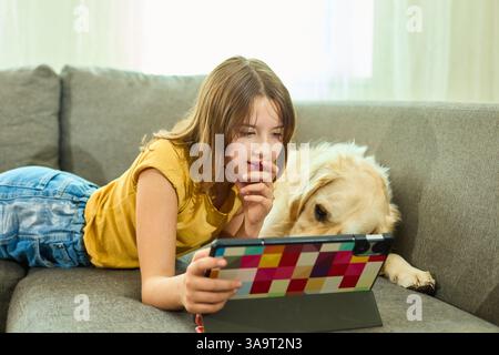 Une adolescente se trouve confortablement sur le canapé dans un salon confortable, s'engageant avec sa tablette. Son Golden retriever se trouve à proximité, ajoutant de la chaleur au rela Banque D'Images