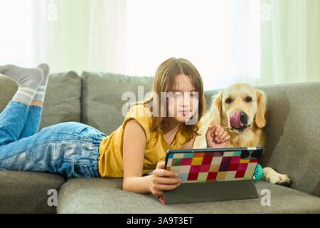 Une adolescente se trouve confortablement sur le canapé dans un salon confortable, s'engageant avec sa tablette. Son Golden retriever se trouve à proximité, ajoutant de la chaleur au rela Banque D'Images