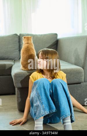 Un moment de calme se déroule dans une maison confortable comme une adolescente assise sur le sol, observant son chat Scottish Fold se reposant confortablement sur le canapé à proximité. Sunlig Banque D'Images