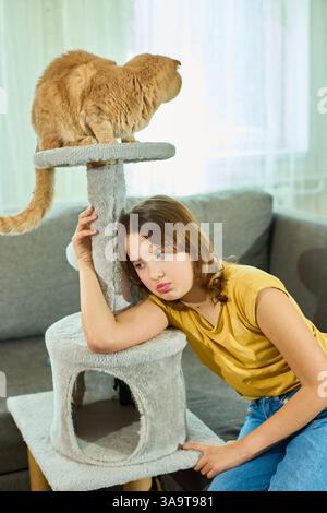 Une adolescente est assise sur le canapé avec une expression ennuyée, reposant sa tête sur un arbre à chat. Son chat Scottish Fold observe les environs depuis le sommet de la ta Banque D'Images