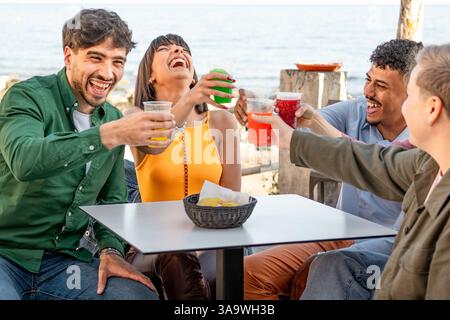 Un groupe de jeunes amis divers riant et grillant avec des boissons colorées dans un café en bord de mer, profitant d'une atmosphère joyeuse et détendue. Banque D'Images