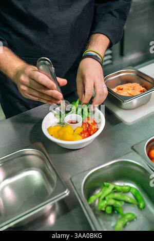 Gros plan sur des mains de chef méconnaissables plaçant des ingrédients avec des pinces dans un bol délicieux et sain poke, comme le saumon, l'edamame, les tomates, la mangue et le s. Banque D'Images