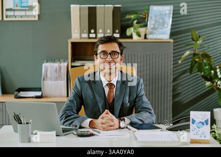 Portrait d'un homme d'affaires portant des lunettes et un costume, assis à un bureau avec un ordinateur portable, des dossiers et des plantes de bureau derrière, souriant et donnant une impression professionnelle Banque D'Images