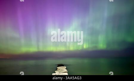 Les aurores boréales à couper le souffle affichent des danses dans le ciel nocturne, mêlant des teintes vertes et rouges vives sur un lac calme avec un rivage rocheux. Banque D'Images