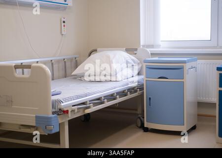 Chambre d'hôpital vide avec lit réglable propre et armoire de chevet bleue près de la fenêtre, linge blanc soigneusement arrangé, environnement stérile lumineux, personnes âgées Banque D'Images