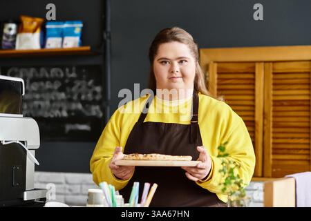 Jeune femme atteinte du syndrome de Down se tient fièrement dans un café, tenant des friandises fraîchement cuites sur un plateau. Banque D'Images