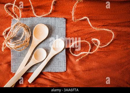 Cuillères en bois sur une serviette grise avec de la ficelle de jute naturelle, disposées sur un fond de tissu rouge texturé. Concept de cuisine rustique et écologique avec guerre Banque D'Images