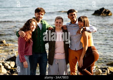Un groupe diversifié d'amis souriant et se liant au bord de la mer par une journée ensoleillée, profitant d'un moment joyeux et détendu ensemble. Banque D'Images