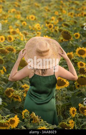 Femme en robe verte debout dans le champ de tournesol, tenant le chapeau jaune dans les mains. Photographie artistique Banque D'Images