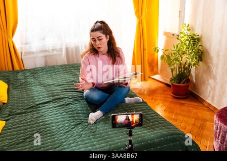 Femme parlante assise sur un lit et vlogging avec un smartphone, créant du contenu de médias sociaux de haute qualité pour ses abonnés en ligne. Grand angle. Banque D'Images