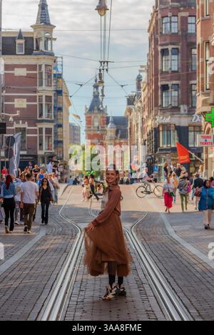 Une femme élégante dans une robe marron fluide marchant en toute confiance sur les voies de tramway d'Amsterdam, mêlant mode et énergie de la ville Banque D'Images