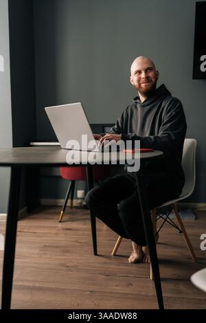 Portrait vertical pleine longueur de programmeur joyeux masculin travaillant à distance du bureau à domicile, assis pieds nus à la table en bois, tapant sur un ordinateur portable Banque D'Images