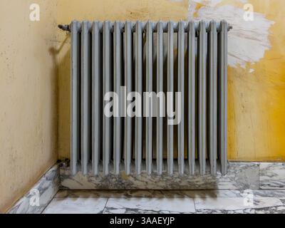 Vieux radiateur en fonte grise sur mur sale avec plâtre jaune sur sol en marbre blanc Banque D'Images