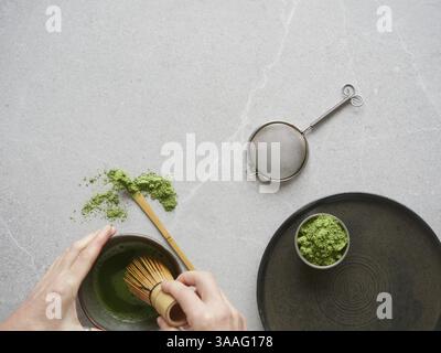 Préparation de boisson au thé vert avec de la poudre de matcha. Mains avec un fouet traditionnel en bambou sur un bol de boisson au thé matcha sur une table en pierre grise Banque D'Images