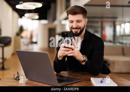 Un jeune homme souriant employé de bureau est assis dans le bureau à un bureau et utilise un téléphone portable, écrit un message, correspond. Banque D'Images
