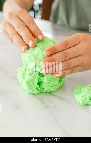 Gros plan des mains d'une personne moulant de la pâte vert vif sur une surface de marbre lisse. L'image capture l'activité tactile de la mise en forme et de la compression Banque D'Images