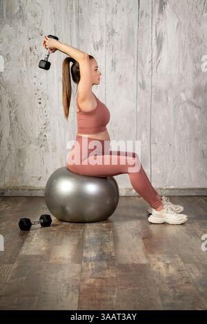 Sportive enceinte exerçant avec des haltères assis sur un ballon de fitness dans un gymnase Banque D'Images