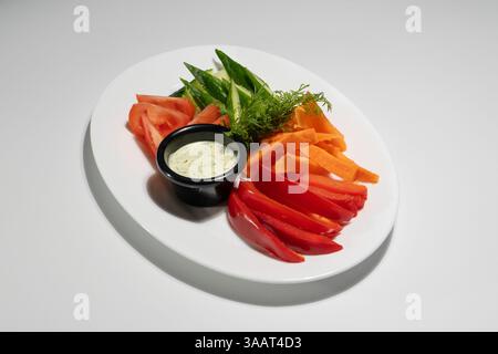 Plateau de légumes frais avec tomates tranchées, concombres, carottes et poivrons servis avec une sauce crémeuse aux herbes sur fond blanc. Banque D'Images