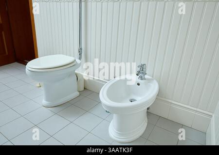 Toilettes blanches et bidet côte à côte dans une salle de bain carrelée. Luminaires de salle de bain classiques contre un mur à panneaux blancs, mettant en valeur une résolution simple et traditionnelle Banque D'Images