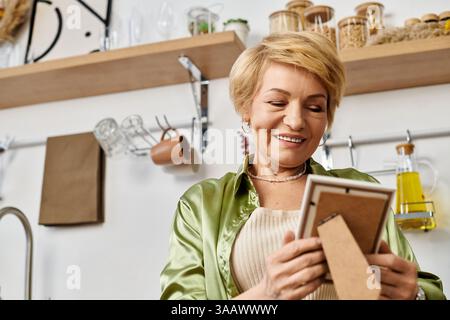 Femme âgée aux cheveux blonds courts sourit, tenant une photo encadrée dans sa cuisine lumineuse. Banque D'Images