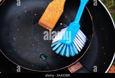 Gros plan d'une poêle sale avec des gouttes de savon liquide, un récureur en bois et une brosse bleue, symbolisant le style de vie du camping, le nettoyage extérieur et la corvée Banque D'Images