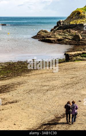 Deux personnes apprécient une promenade tranquille le long d'une plage de Cornouailles, la côte rocheuse et la mer calme offrant une toile de fond tranquille. Banque D'Images