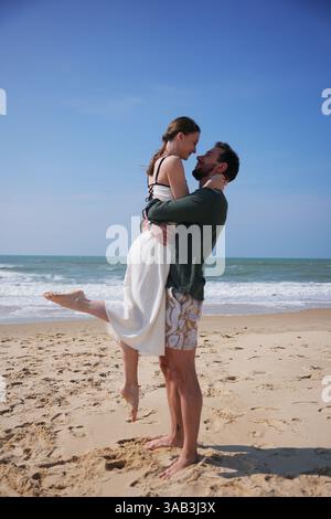 Jeune couple partage une étreinte tendre et joyeuse sur une plage ensoleillée, avec la femme soulevée du sol dans l'affection. Le ciel clair et les vagues douces de l'océan créent un cadre serein d'été plein d'amour Banque D'Images