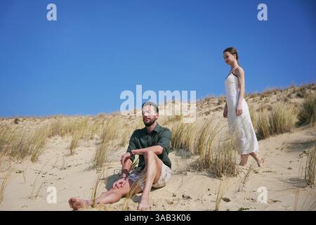 Jeune homme est assis paisiblement sur des dunes ensoleillées tandis qu'une femme en robe blanche marche pieds nus à proximité sous un ciel bleu vif. Banque D'Images