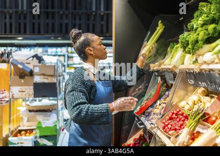 Employé de supermarché portant des gants arrangeant le céleri biologique sur une étagère, entouré de divers produits frais dans une épicerie Banque D'Images