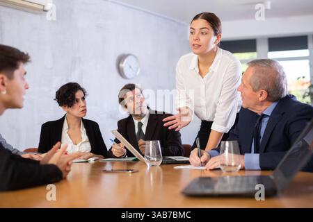 Intelligent jeune femme faisant rapport tandis que les employés de bureau d'âges différents utilisant des cahiers à la table dans le bureau Banque D'Images