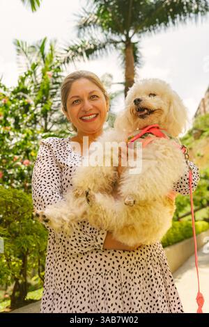 Heureuse femme mature tenant et câlinant son caniche blanc moelleux tout en marchant dans un parc ensoleillé, profitant d'une belle journée à l'extérieur avec son bien-aimé Banque D'Images