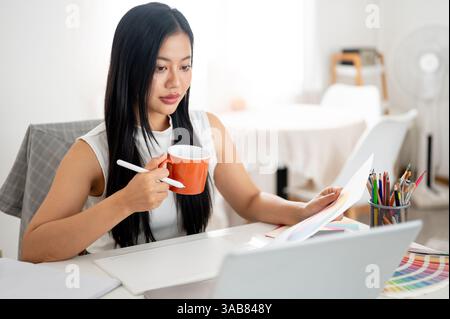 Une femme asiatique tient une tasse en céramique orange avec un stylet tout en regardant l'écran de l'ordinateur portable. Designer d'intérieur ou Graphic Designer au bureau. Banque D'Images