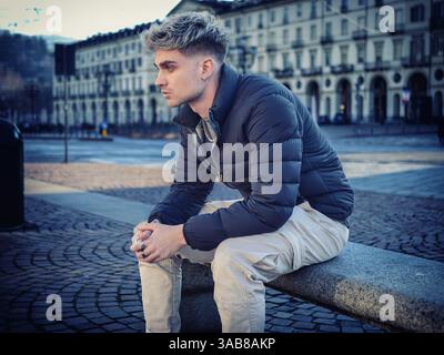 Un jeune homme aux caractéristiques attrayantes est assis pensivement sur un banc de pierre dans un cadre urbain calme. Ses cheveux blonds et ses yeux bleus se détachent sur fond de bâtiments en plein jour. Banque D'Images