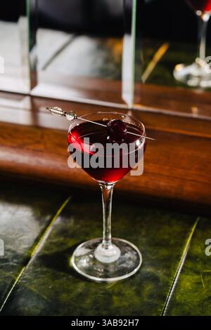 Un cocktail de gin riche aux fruits rouges avec des notes florales et des éclaboussures de vin mousseux, servi dans un élégant verre coupé. La garniture de cerise foncée et miroir Banque D'Images