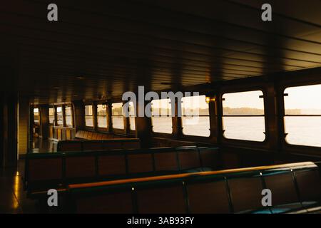Intérieur du ferry vide au coucher du soleil, avec bancs, fenêtres et vue sur l'eau sereine à New York. Un éclairage chaud crée un cadre paisible et réfléchissant Banque D'Images