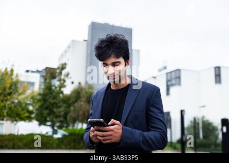 Un homme d'affaires indien en costume de marine, tenant un smartphone, se tient dehors, concentré sur son appareil. Illustrer une ambiance professionnelle lors d'une conférence de Banque D'Images