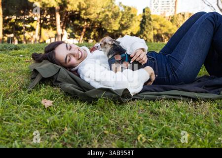 Une femme en pull blanc se trouve dans le parc, souriante et tenant un petit chihuahua. La lumière du soleil filtre à travers les arbres, créant une scène extérieure sereine. Banque D'Images