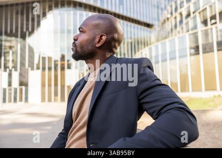 Un homme d'affaires cubain réfléchi dans un costume de marine regarde au loin, debout devant un immeuble de bureaux en verre moderne, symbolisant l'ambition et Banque D'Images