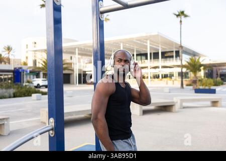 Un homme cubain dans un débardeur noir et un short gris écoute de la musique sur des écouteurs. Il se tient à l'extérieur près d'un bâtiment moderne, détendu et concentré, sous Banque D'Images