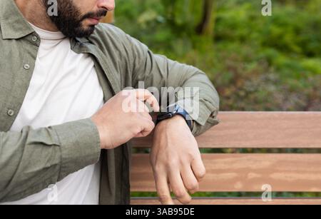 Un homme interagit avec sa smartwatch tout en étant assis sur un banc en bois dans un cadre luxuriant dans la ville. Il porte une chemise verte décontractée sur un tee-shirt blanc, se concentrant sur Banque D'Images