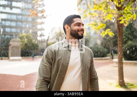 Un homme à la barbe sourit tout en profitant d'une légère pluie dans un parc de la ville. Il porte une veste légère, entourée d'arbres et de bâtiments modernes, captur Banque D'Images
