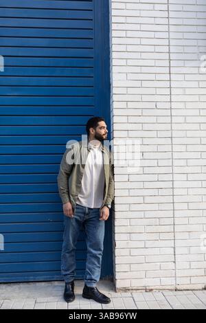 Un homme en tenue décontractée s'appuie contre un mur de briques blanches de la ville, avec une porte bleue à côté de lui. Il porte un Jean, un T-shirt blanc et un ja vert Banque D'Images