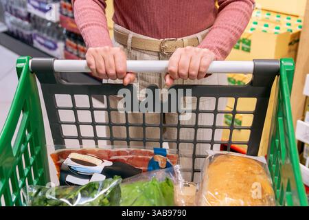 Une image d'une femme poussant un chariot rempli de divers produits d'épicerie, y compris du pain et des légumes verts, dans une allée de supermarché. L'accent est mis sur les han Banque D'Images