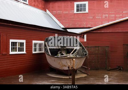 Un vieux bateau altéré est exposé contre un bâtiment en bois rouge vibrant à Hoonah, Alaska, reflétant le patrimoine maritime de la comm Tlingit Banque D'Images