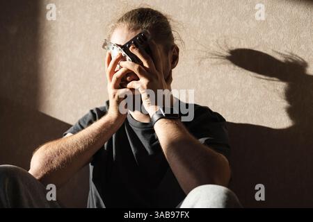 Un homme est assis dans une pièce faiblement éclairée tenant un blister paquet de pilules, profondément dans la pensée. L'ombre subtile sur le mur souligne son humeur pensive et introsp Banque D'Images