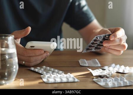 Un homme tient un paquet de pilules dans une main et un smartphone dans l'autre, assis à une table en bois avec des blisters dispersés et un verre d'eau. Banque D'Images